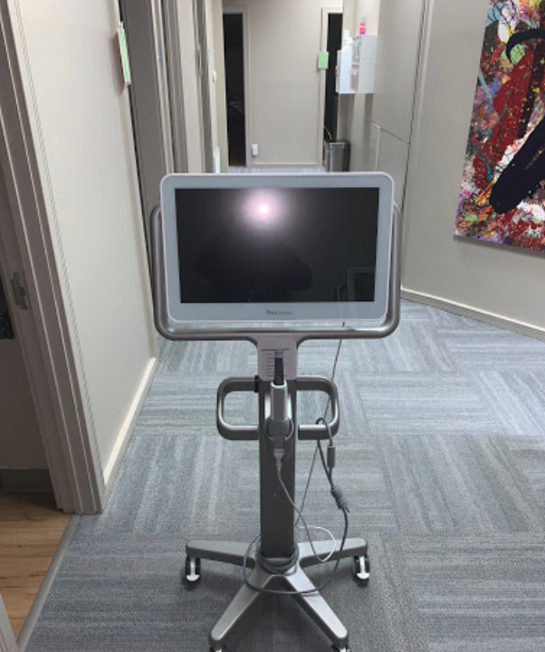The image shows a standing tablet on a mobile stand with wheels, placed on a carpeted floor inside an office space with modern furnishings.