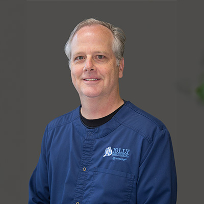 The image shows a person posing for a portrait, dressed in professional attire which includes a blue shirt with an emblem on the left side of the chest, likely indicating a medical profession, and a name tag that reads  Dolly .