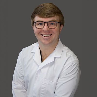 A man wearing glasses and a white lab coat stands smiling against a neutral background.