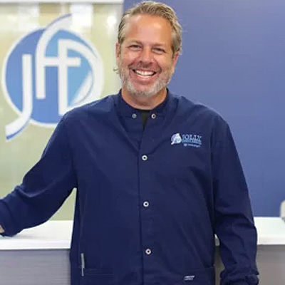 The image shows a man wearing an apron standing behind a counter with a logo in the background, smiling at the camera.