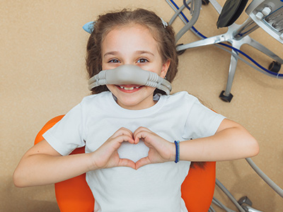 The image shows a young girl sitting on an orange chair with her hands placed over her heart, wearing a medical device that appears to be a nasal oxygen cannula, smiling at the camera.