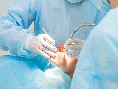 The image shows a medical setting with a person wearing protective blue scrubs and a surgical mask, performing a procedure on another individual who is lying down and has their eyes closed.