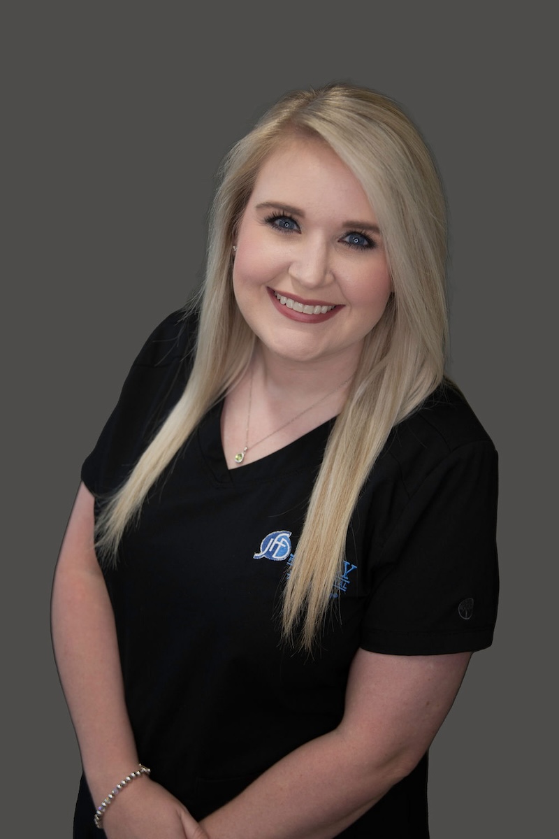 A woman with blonde hair wearing a black polo shirt with a logo on the left side, posing for a professional portrait against a plain background.