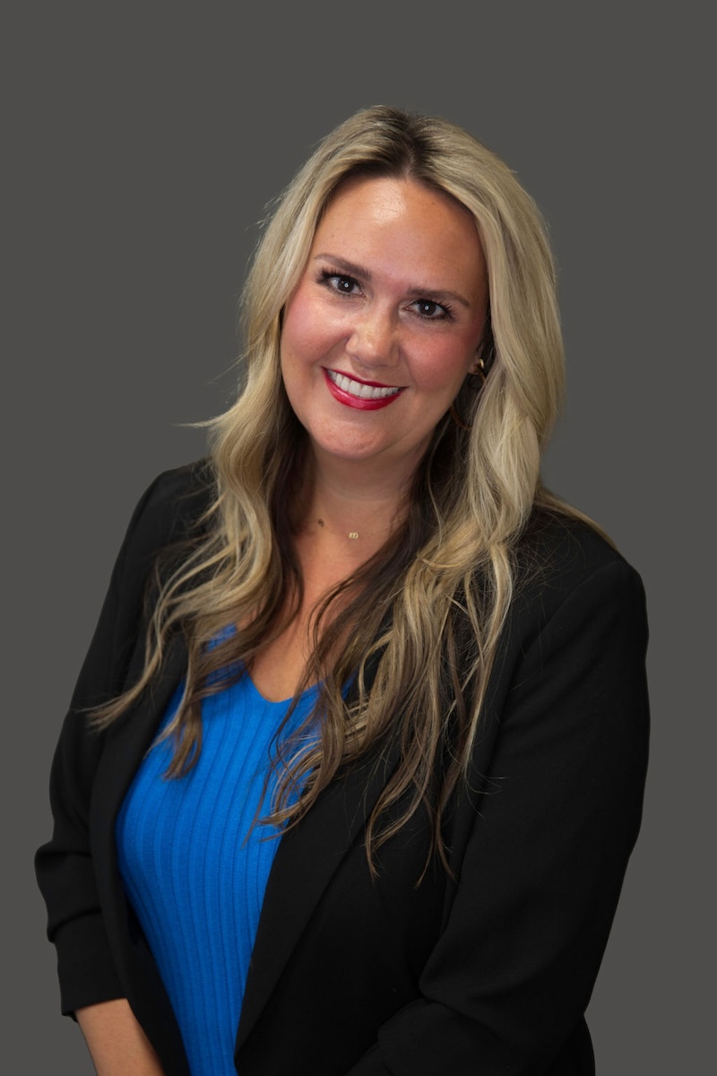 The image shows a woman standing against a neutral background, wearing a dark blazer over a lighter garment with a blue top underneath, smiling at the camera with her hair pulled back into a ponytail.