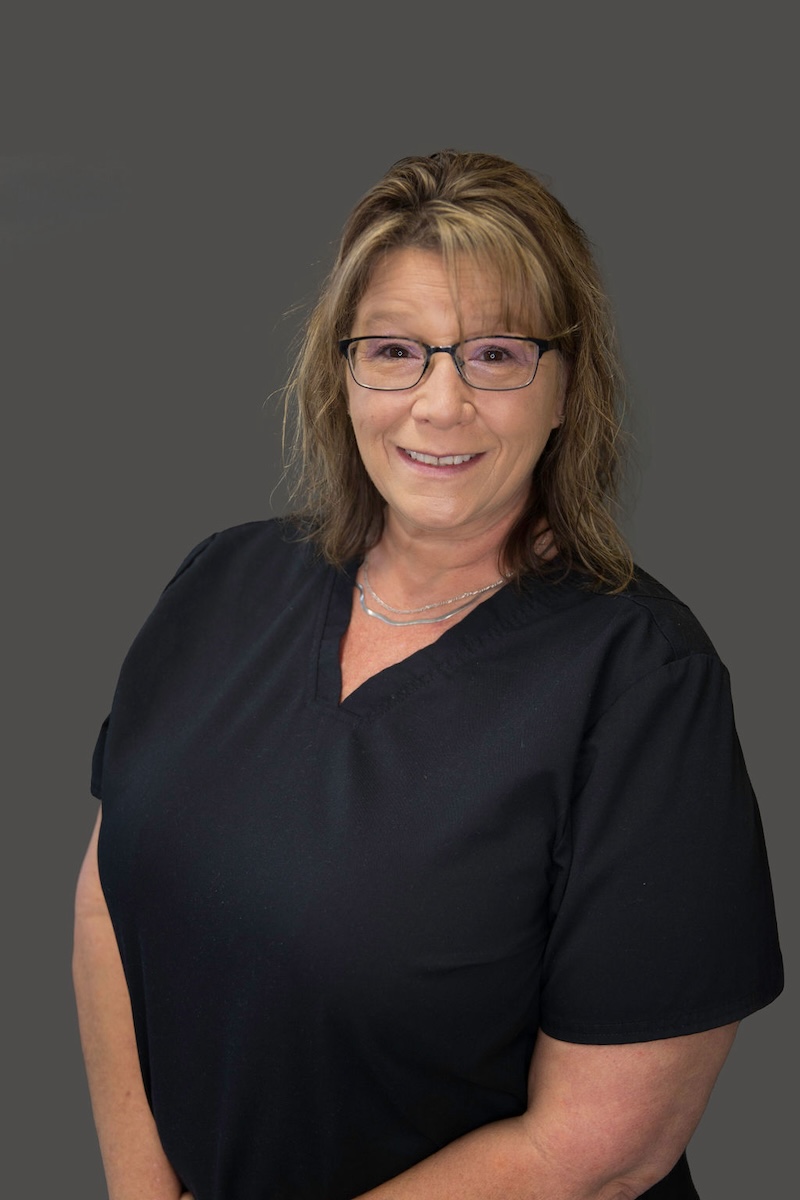 The image displays a woman standing against a neutral background, wearing a dark top with a light-colored collar, glasses, and a name tag, posing for the photograph.