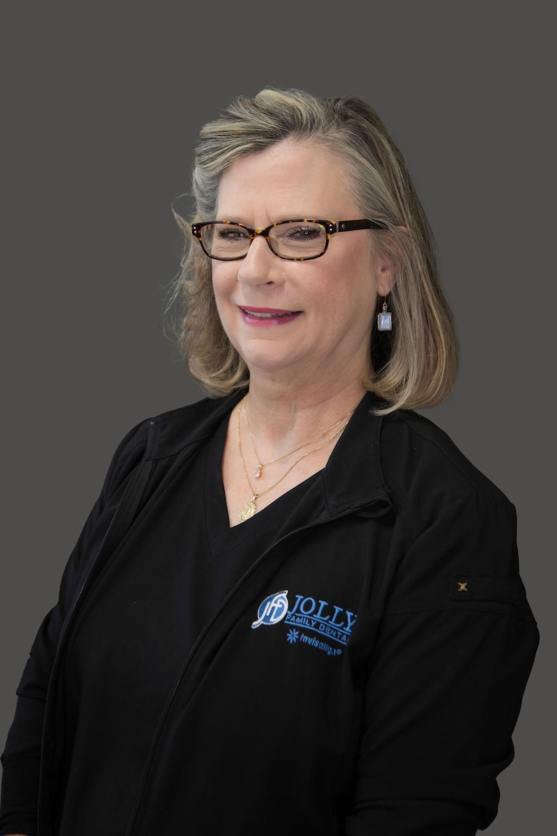 The image shows a woman wearing glasses, with short hair, posing for a professional portrait against a neutral background. She is dressed in a dark top and has a name tag that reads  Jolly.