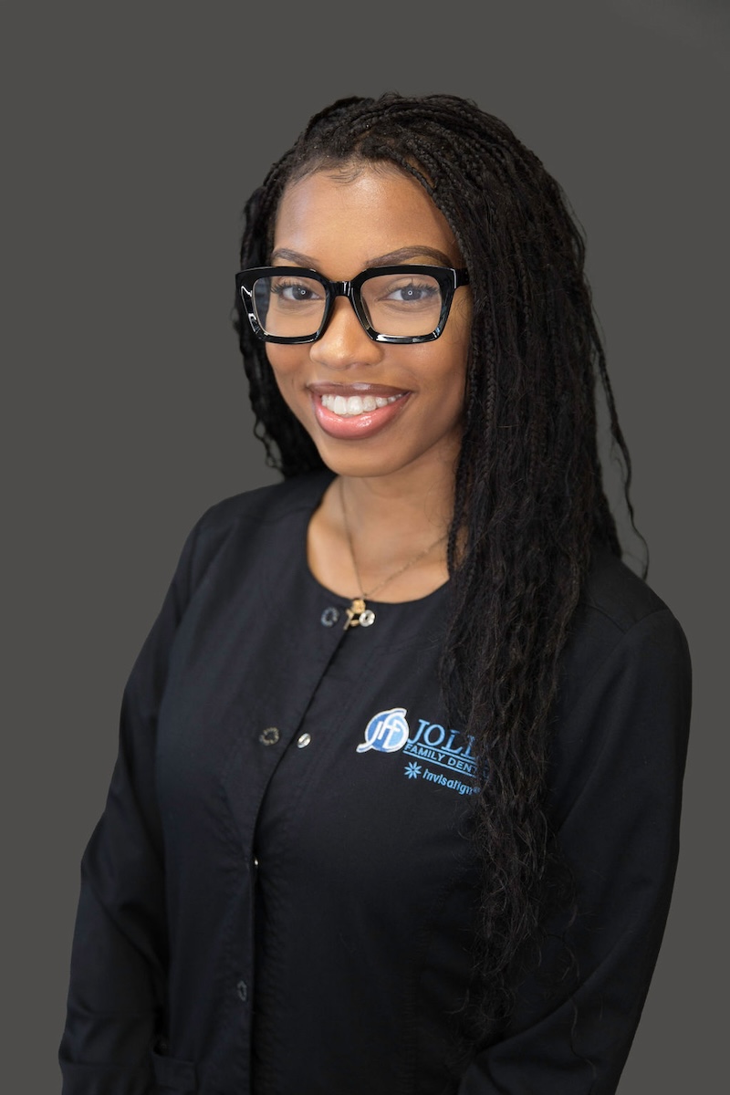 The image shows a woman posing for a professional portrait with a smile, wearing glasses, a black top, and standing against a neutral background.
