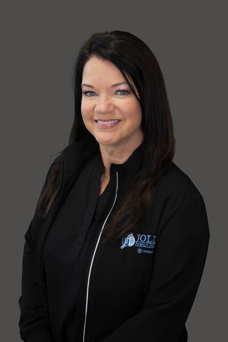 The image shows a woman with long hair smiling at the camera. She is wearing a black jacket with a logo on the left chest area, suggesting she might be a professional or part of an organization.