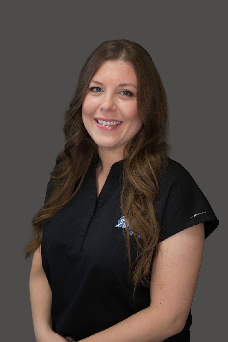 The image shows a woman standing against a gray background. She has long brown hair, is wearing a dark-colored shirt with short sleeves, and has a name tag on her attire. Her expression is neutral, and she appears to be posing for the photograph.