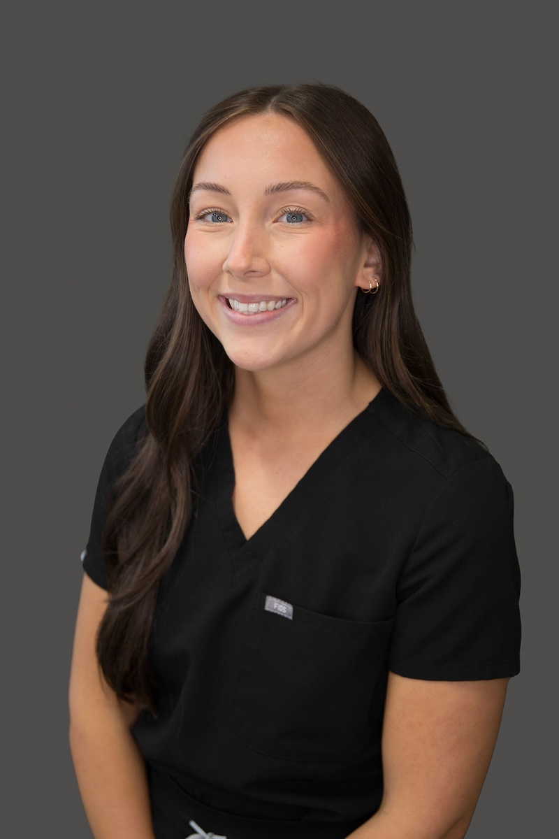 The image shows a woman wearing a black scrub top with a name tag, standing against a neutral background.