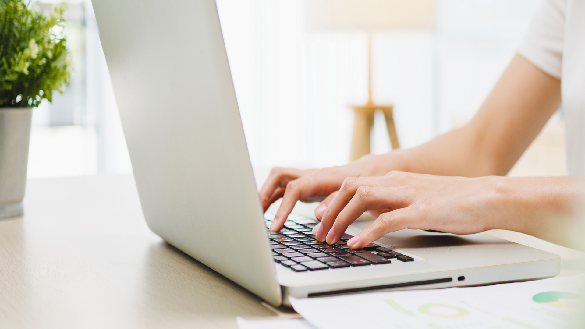 A person's hands typing on a laptop keyboard with a blurred background featuring a plant and a portion of a desk.