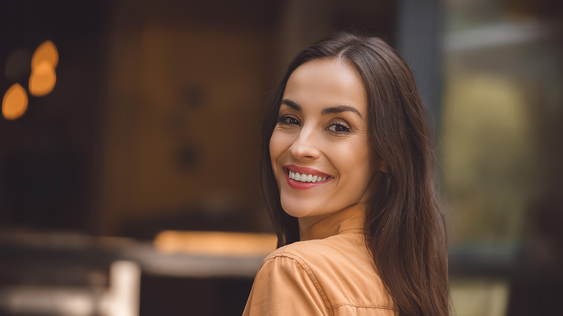 A smiling woman with dark hair wearing a brown top poses in front of a blurred indoor setting.