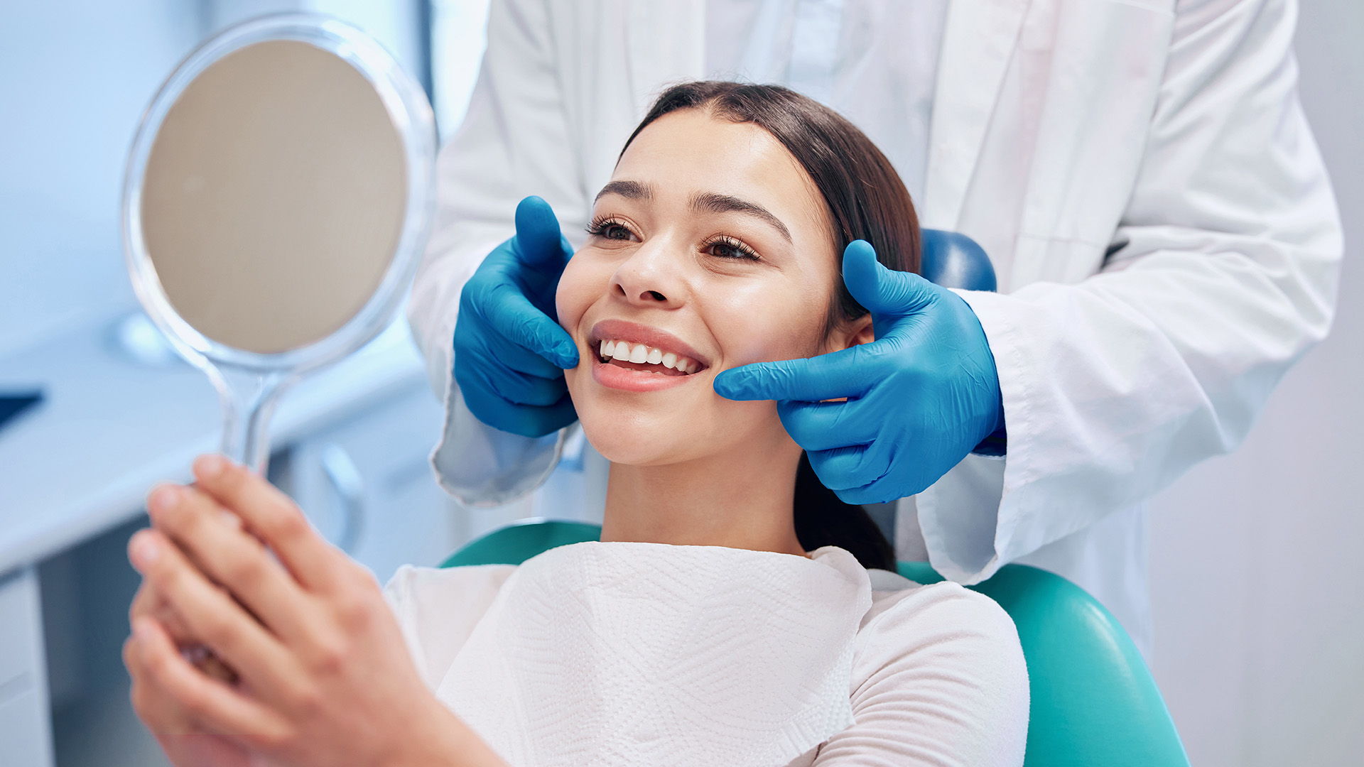 A woman sitting in a dental chair receiving teeth cleaning service from a dentist wearing blue gloves and a white coat.