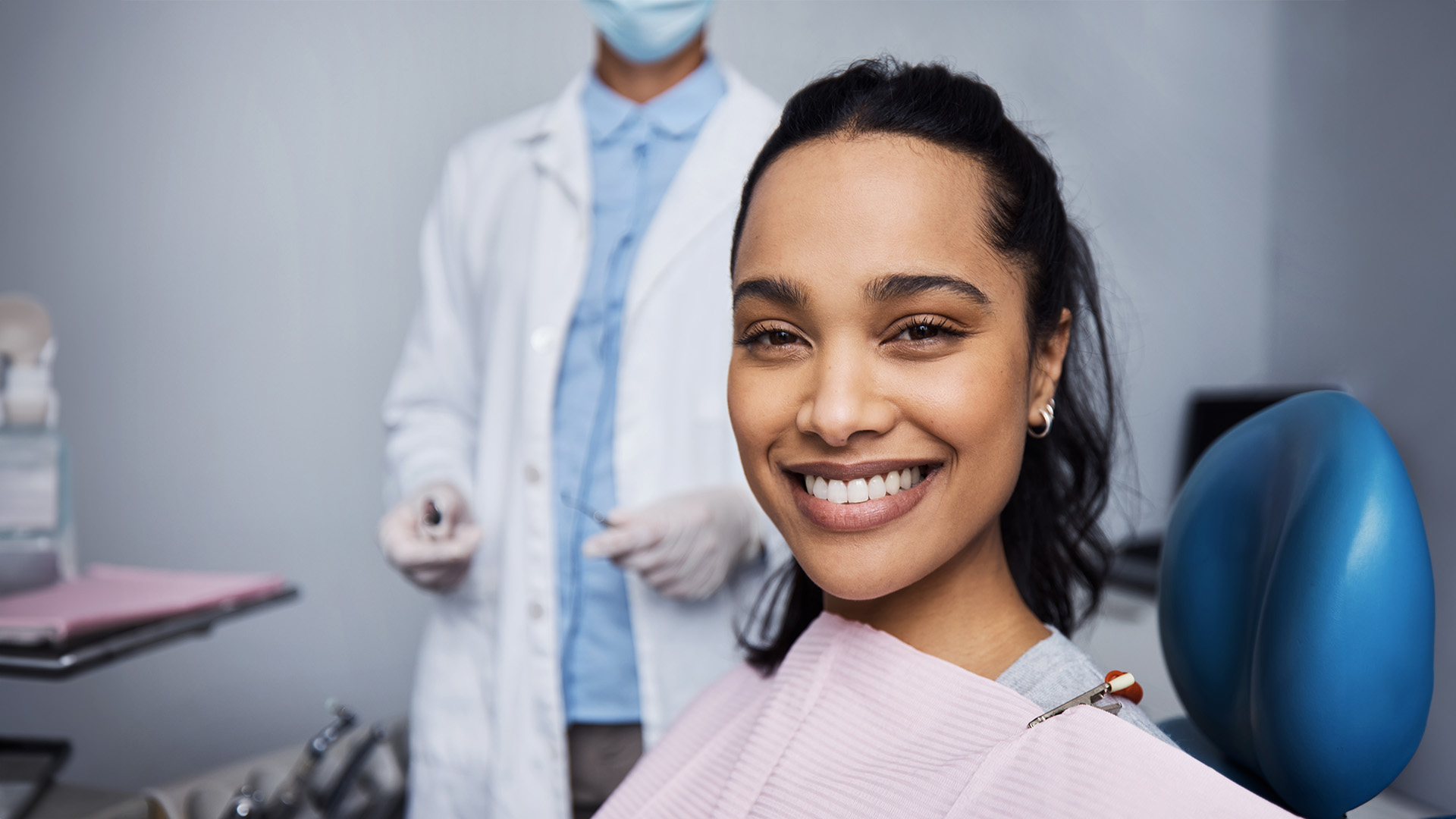 A woman sitting in a dental chair with a smile on her face, receiving dental care from a professional wearing protective eyewear and a white coat.