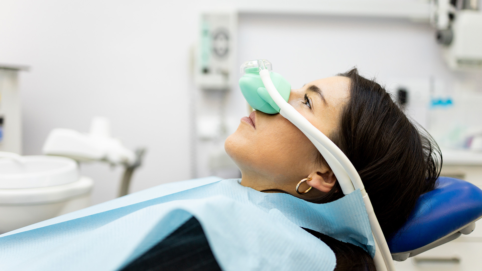 A woman wearing a medical mask over her mouth, sitting in a dental chair with a dental hygienist s headrest, receiving dental treatment in a dental office setting.