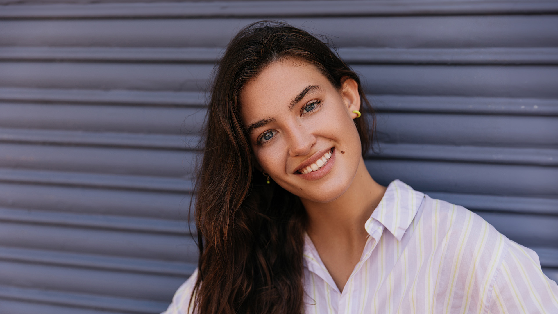The image shows a smiling woman with long hair leaning against a blue wall, wearing a white top and gold earrings.