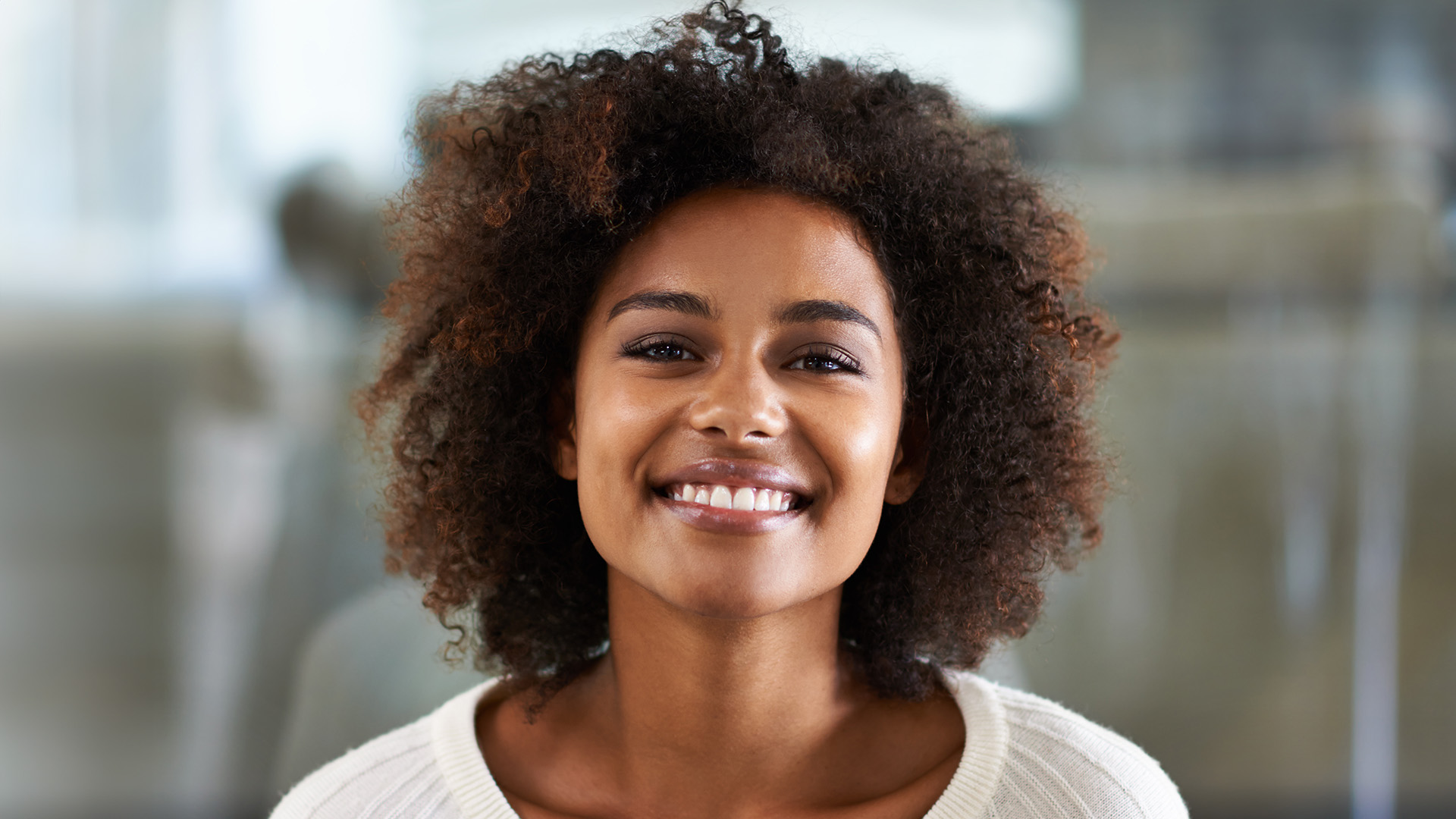 A smiling woman with curly hair looking directly at the camera, set against a blurred background of an office space.