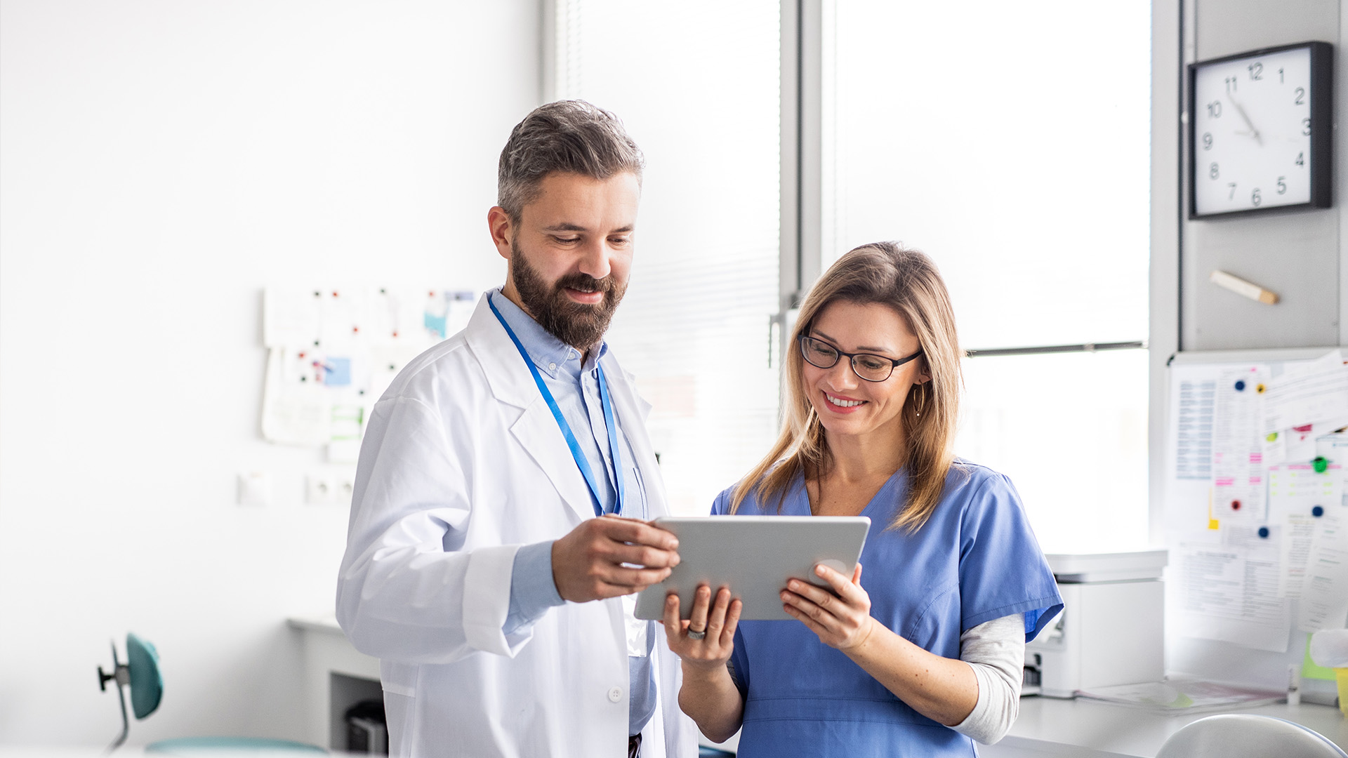 The image shows a man and woman in a medical setting, with the man holding a tablet and both looking at it, while the woman appears to be taking notes.