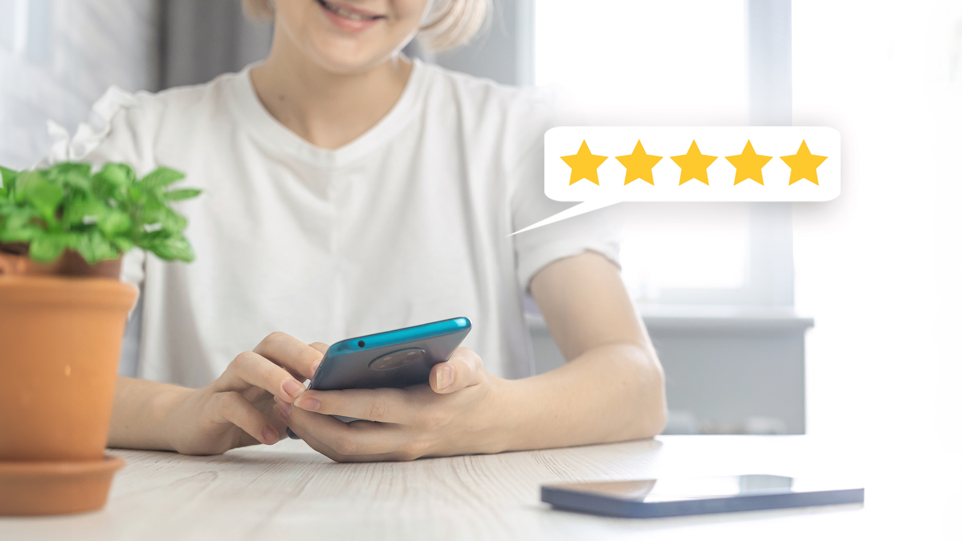 The image shows a woman sitting at a table with a smartphone in her hands, smiling at the camera while looking up from the device. She appears to be engaged in an activity on the phone, which has a five-star rating icon visible on its screen.