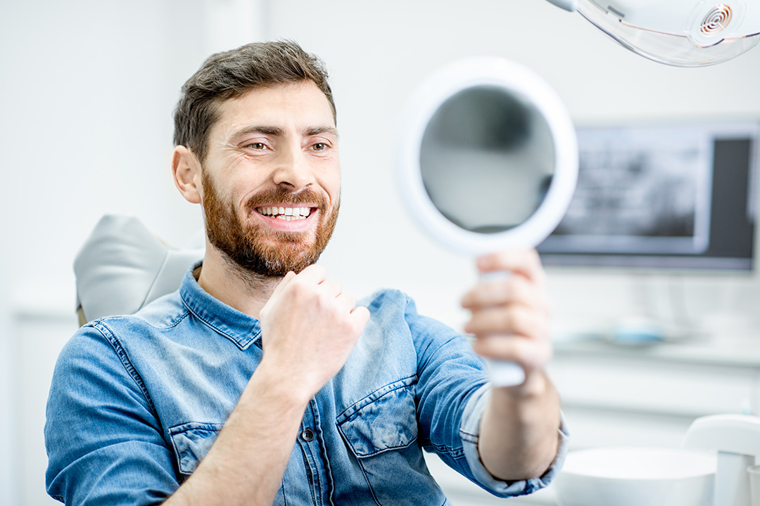 The image features a man with facial hair, smiling broadly, holding up a magnifying mirror while sitting in front of a dental chair with various dental instruments around him.