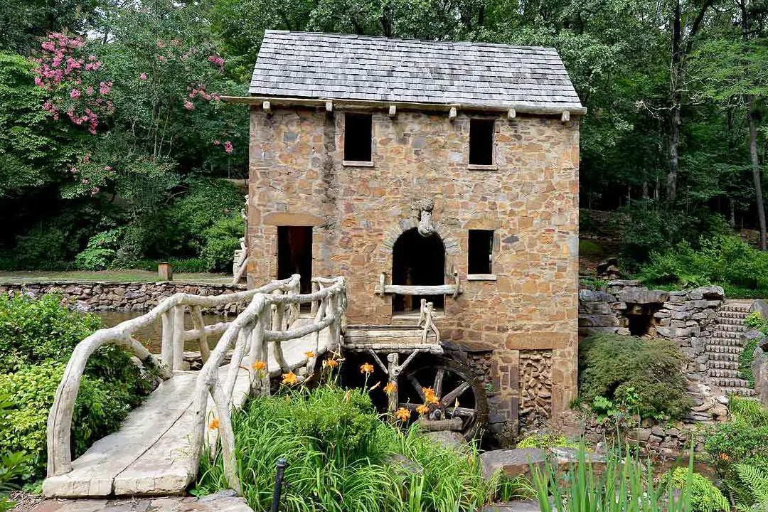 The image shows a quaint stone cottage with a small wooden bridge leading up to it, surrounded by lush greenery and a tranquil forest setting.