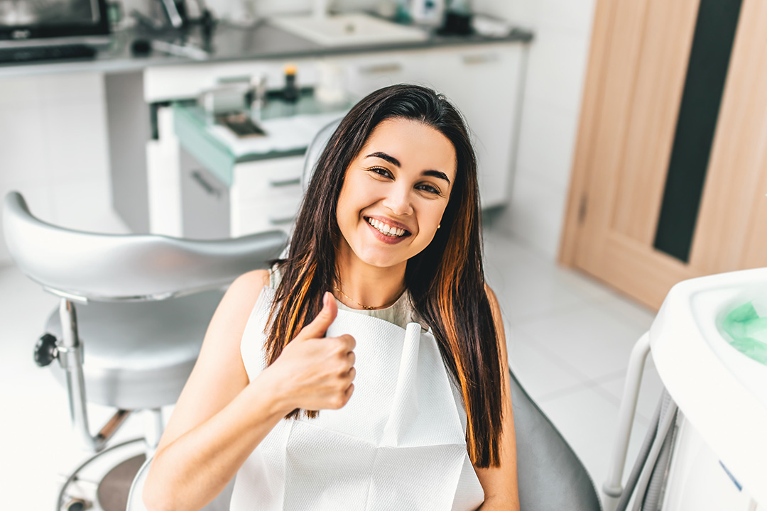 A young woman giving a thumbs-up gesture while sitting in a dental chair with a dental hygienist s room in the background.