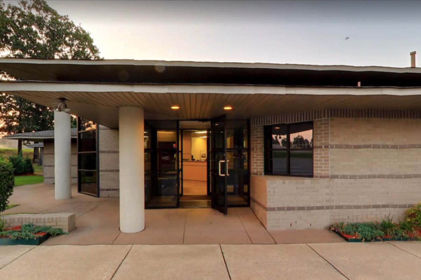 The image depicts an exterior view of a single-story building with architectural features such as a flat roof, overhanging eaves, and a prominent central entrance. The building appears to be a public facility, possibly a community center or a library, indicated by its modern design and the presence of what looks like a sign on the wall above the entrance.