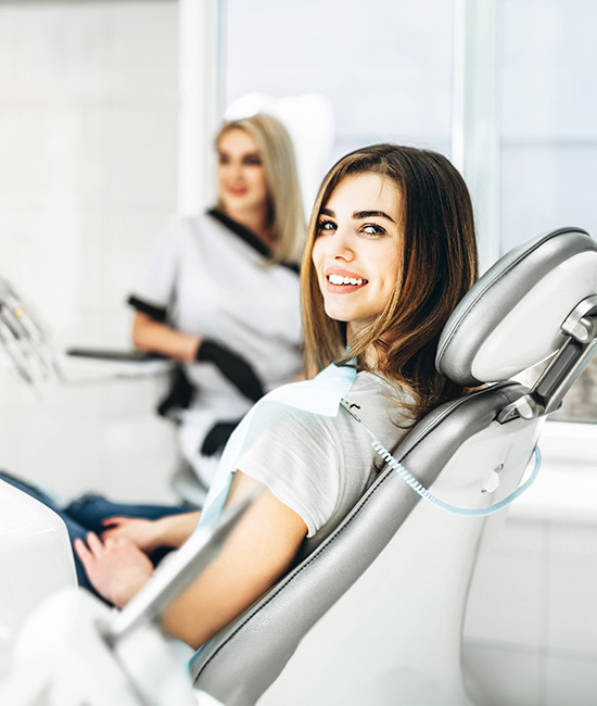 A woman sitting in a dental chair with a smile, next to a female dental professional in a white coat and stethoscope.