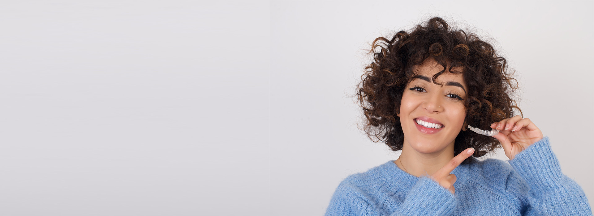 A woman with short hair smiles at the camera while holding a phone near her face. She has curly hair and is wearing a blue sweater.