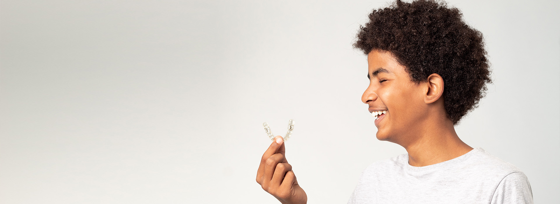 A young man with a beard, wearing a white t-shirt, smiling broadly while holding a seedling, against a plain background.