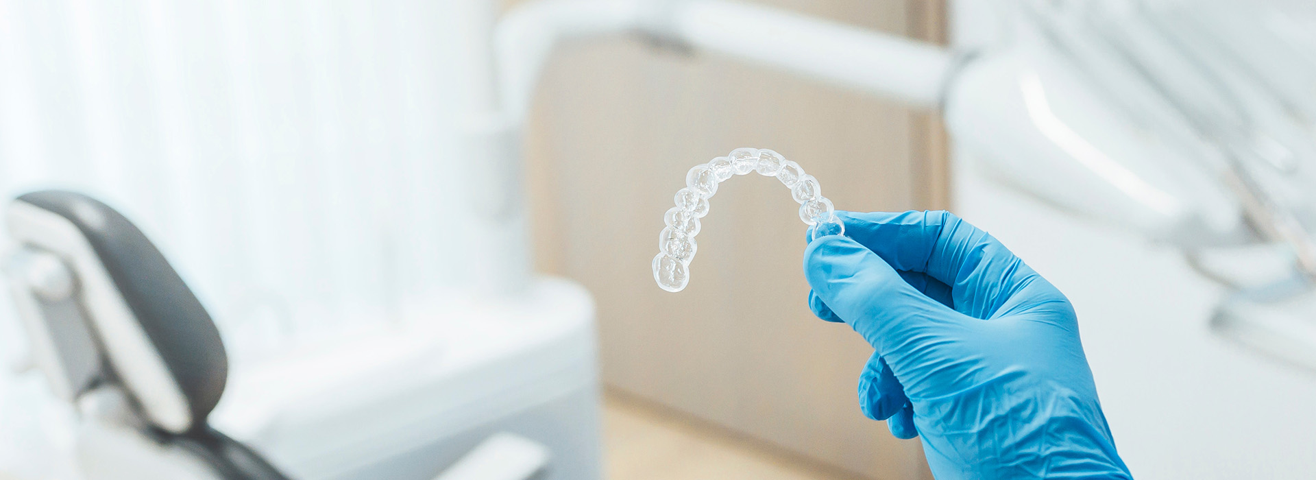 The image shows a close-up of a transparent dental impression being held by a hand wearing blue gloves, with a blurred background featuring a dental office setting.