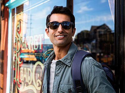 Man standing outdoors, smiling at camera, wearing sunglasses, jacket, and backpack, positioned in front of a shop window with a sign.