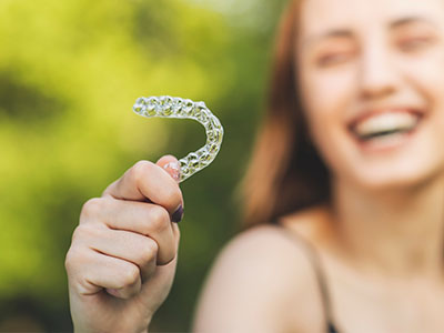 The image features a young woman holding up a clear plastic retainer with her left hand while smiling at the camera.