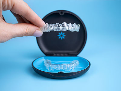 This is an image of a person s hand holding a transparent dental impression tray with a blue base, which is placed on a black box containing clear plastic teeth aligners.