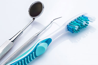The image displays a collection of dental tools including a toothbrush with blue bristles, a pair of scissors, and a small spoon-like instrument on a light background, suggesting a dental care setting.