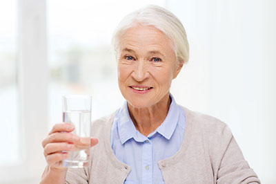 The image features an elderly woman holding a glass of water with her left hand while smiling at the camera.