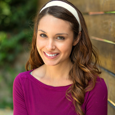 A smiling woman with long brown hair, wearing a purple top and headband, posing against a wooden fence.
