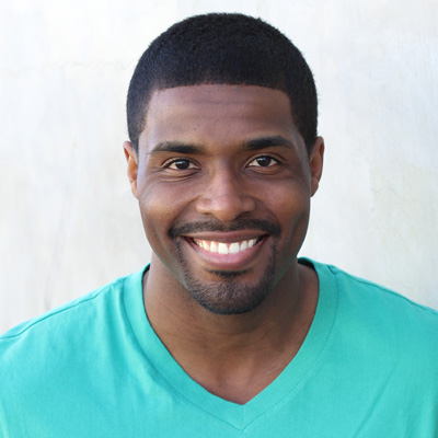 An elderly couple smiling at the camera, with the man wearing a gray beard and mustache, standing close together outdoors during daylight.
