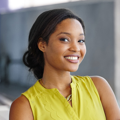 The image features a smiling woman with dark hair, wearing a yellow top, posing against a blurred background that includes a building structure.