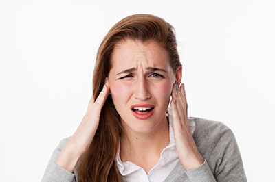 The image shows a young woman with her hand on her ear, appearing to be experiencing discomfort or pain, possibly due to an issue with her hearing. She has blonde hair and is wearing a gray sweater.