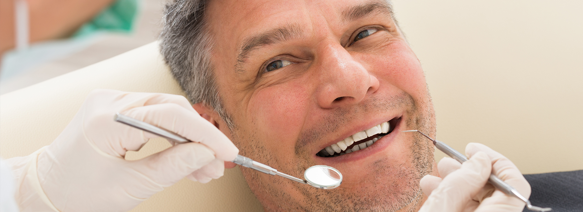 The image shows a man seated in a dental chair with his mouth open, receiving dental care from a practitioner who is working on him while wearing gloves and surgical tools.