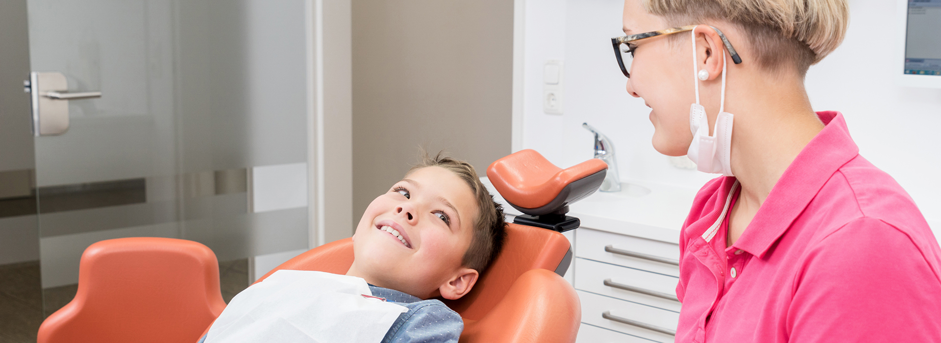A young boy seated in a dental chair with a woman standing behind him, both in a dental office setting.