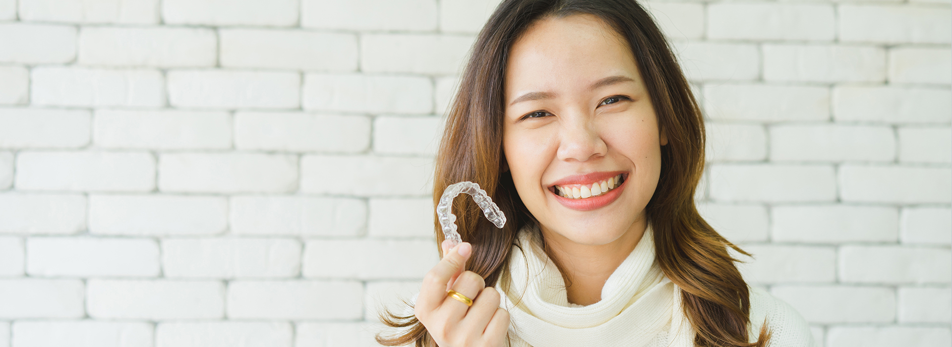 A smiling woman wearing a white top holds a ring with both hands against a brick wall background.