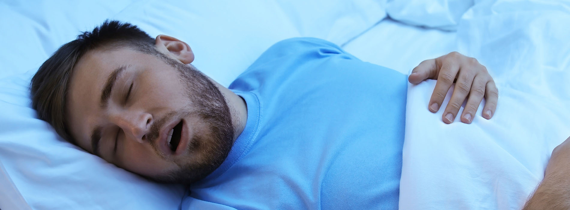 The image shows a man lying down in bed with his eyes closed, wearing a blue shirt and a white mask covering his face.