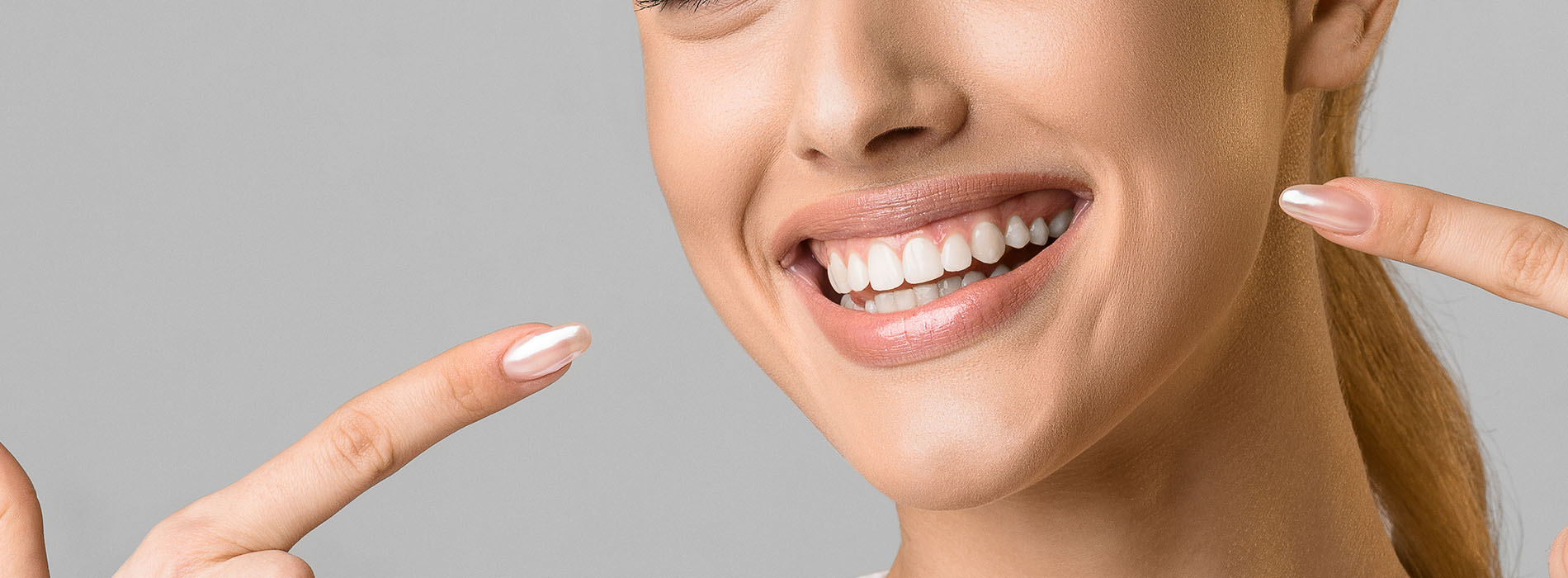 The image shows a smiling woman with her eyes closed, holding up two fingers on each hand near her face, against a plain background.