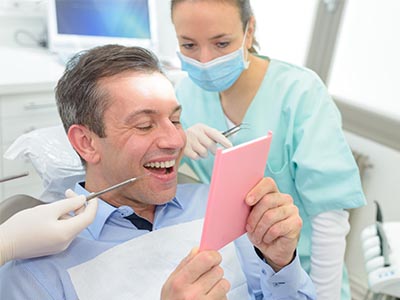 The image shows a man sitting in a dental chair with a big smile on his face, holding up a pink card, while a dental professional stands behind him, both appearing to be in a dental office setting.