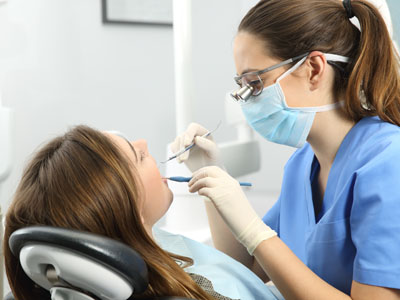 The image shows a dental hygienist performing a teeth cleaning on a patient while wearing personal protective equipment.