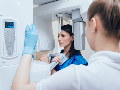 A woman in a blue coat stands next to a large, white 3D scanner with a digital display, while another person is seated in front of it wearing gloves and a face mask.