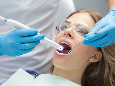 A woman receiving dental treatment with a dental device inserted into her mouth by a dental professional wearing protective gloves and a face mask, while she sits in a dental chair.