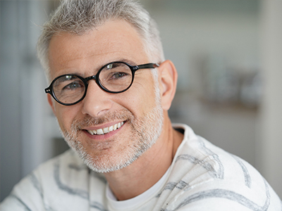 The image features a man with short hair, wearing glasses, smiling at the camera with his head slightly tilted. He has a beard and mustache, and he appears to be indoors, possibly in an office setting, given the presence of a blurred background that suggests a professional environment.